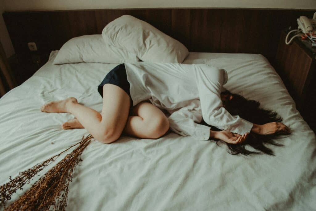 一位長髮女性穿著白色襯衫和深色短褲,居家清潔後疲憊地側躺在白色床單上的深色床頭板前,旁邊有乾燥的植物枝條。