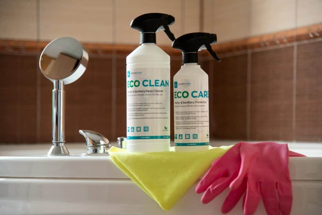 在浴缸邊緣放置著兩瓶清潔產品，分別標示為 ECO CLEAN 與 ECO CARE，前方有一條黃色清潔布與一雙粉紅色橡膠手套，背景為棕色調的浴室瓷磚。