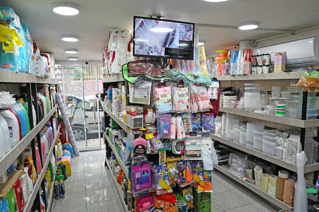 一間擺滿貨物的小型雜貨店走道，左側貨架排滿了五顏六色的洗衣精與清潔劑瓶罐，右側則有塑膠保鮮盒與免洗餐具，走道上方掛著監視器螢幕。