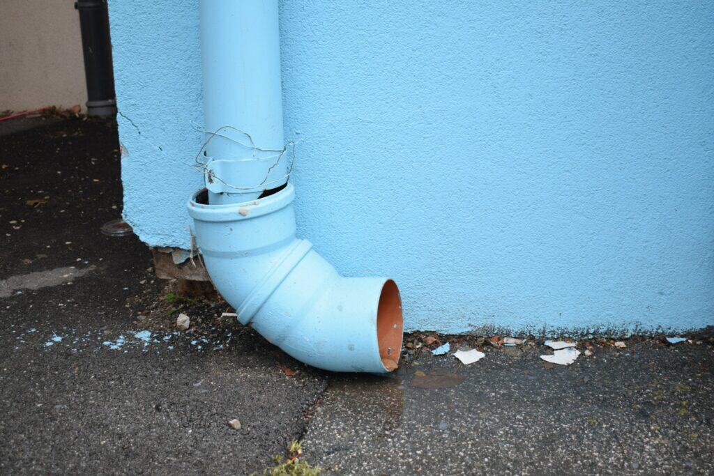 天藍色牆面背景，地面連接處有一根淺藍色 PVC 排水彎管，管道邊緣有鐵絲纏繞加固痕跡，地面可見少許水漬。