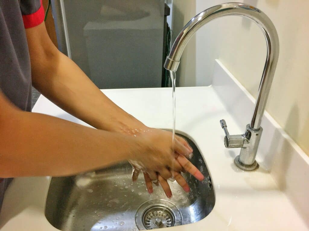 在白色陶瓷洗手台中，水龍頭正流出清水，一名使用者正在洗手，下方排水口處清晰可見，展示日常用水環境。