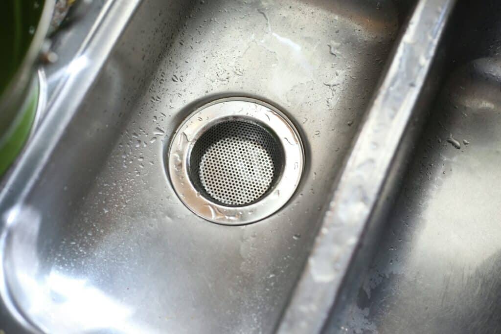 近距離拍攝不鏽鋼廚房水槽，排水口中心配有細密的小孔過濾網，水槽表面帶有少許水滴，整體呈現乾淨的金屬光澤。