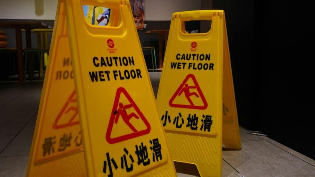 兩座黃色的 A 字型清潔警告牌，上面印有「CAUTION WET FLOOR」與「小心地滑」的中英文字樣及滑倒的人形圖示，背景為商場或公共區域。