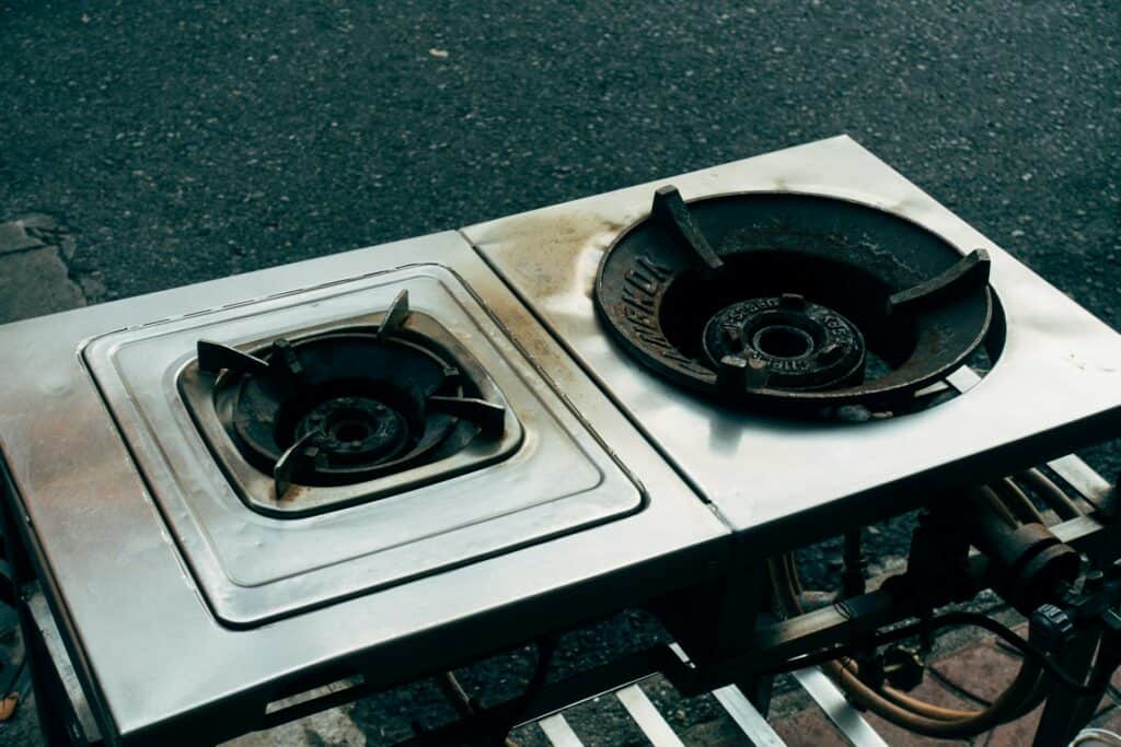 一台放置在室外地面的工業用雙頭不鏽鋼瓦斯爐具，爐口帶有黑色鑄鐵支架，金屬表面有使用過的痕跡與輕微油漬。