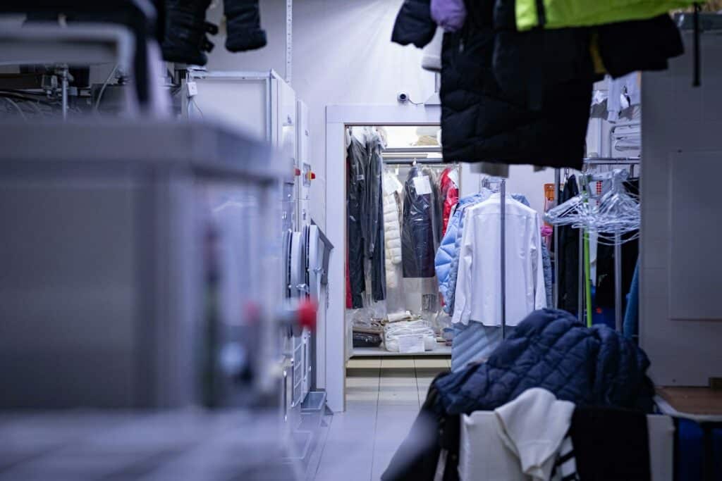 洗衣店或工廠內部的長廊視角，兩側掛滿了洗淨並套上塑膠防塵套的西裝、襯衫與外套，遠處有工作區域的白色櫃體與照明。