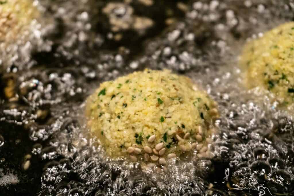 近距離拍攝油鍋中正在油炸灑有芝麻的淺色蔬菜丸子，油花沸騰噴濺，充滿油脂感。