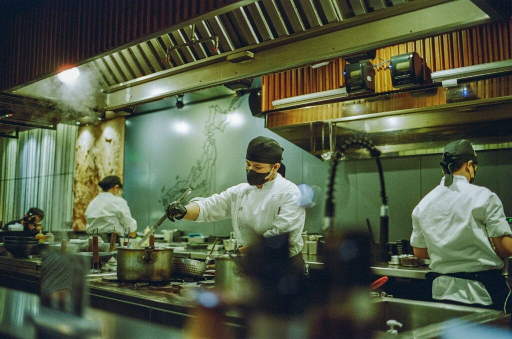 營業用專業廚房的實際工作場景，多位穿著白色廚師服、戴著黑色帽子與口罩的廚師正在不鏽鋼爐台前忙碌烹飪。爐火上放著大型湯鍋，畫面中瀰漫著熱氣與水蒸氣，上方為大型金屬排油煙罩。