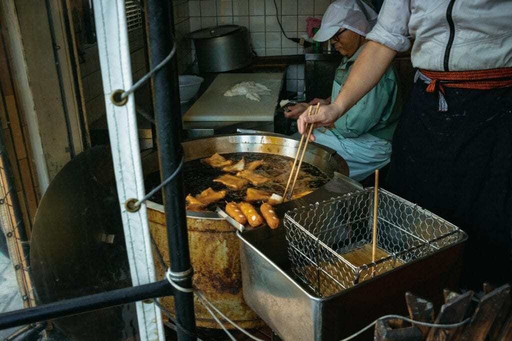 攤販正在炸物的畫面,大油鍋內翻滾著炸食,鍋緣佈滿深色焦油垢,工作人員戴著帽子與圍裙正在操作,強調重油污的使用背景。